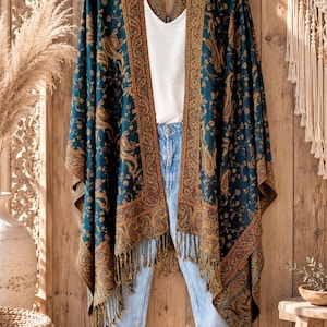 May include: A teal kimono featuring a dark orange and gold paisley pattern with a wide border, open front, and fringe trim along the bottom hem, layered over a white t-shirt and light blue denim jeans on a black hanger.