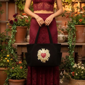 Könnte beinhalten: Schwarze Tragetasche mit goldenem und rotem Herzdesign. Die Tasche hat zwei Außentaschen und schwarze Griffe. Die Tasche wird vor einer Person gehalten, die ein bordeauxrotes Spitzen-Top und einen Rock trägt.