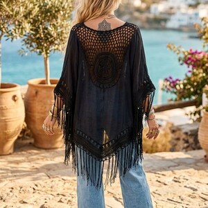 K&ouml;nnte beinhalten: Ein schwarzer Poncho mit einem geh&auml;kelten R&uuml;ckenteil mit kreisf&ouml;rmigem Muster, einer Vorderseite aus festem Stoff und langen Fransen am Saum und an den &Auml;rmeln. Das Kleidungsst&uuml;ck wird &uuml;ber einer hellblauen Jeans getragen und wirkt strukturiert.