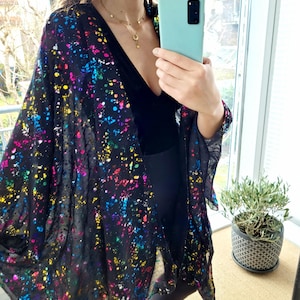Könnte beinhalten: Schwarzer, durchsichtiger Kimono mit einem farbenfrohen Farbspritzer-Design. Der offen geschnittene Kimono hat eine fließende Silhouette und weite Ärmel. Die lebendigen Farbspritzer umfassen Rosa, Gelb, Blau und Grün. Der Kimono wird über einem schwarzen Outfit getragen.