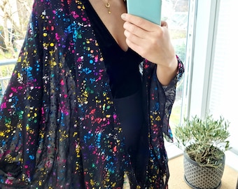 May include: Black sheer kimono with a colorful paint splatter design. The open-front kimono features a flowing silhouette and wide sleeves. The vibrant splashes of color include pink, yellow, blue, and green. The kimono is worn over a black outfit.