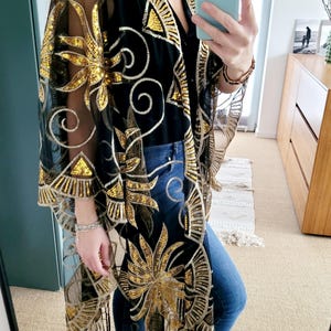 May include: A black sheer kimono featuring intricate gold embroidery and shimmering gold sequins in a floral and geometric pattern. The garment has an open front, wide sleeves, and a hemline finished with long black fringe detailing.