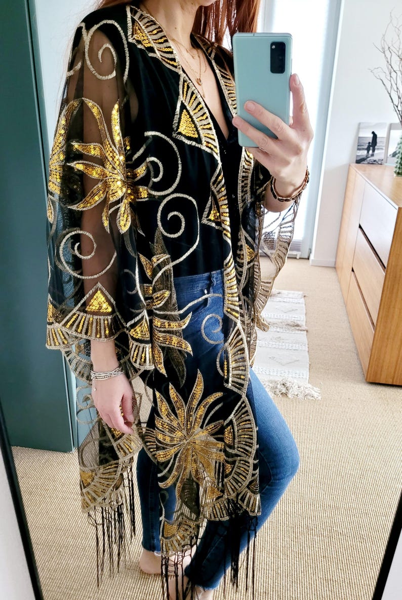 May include: A black sheer kimono featuring intricate gold embroidery and shimmering gold sequins in a floral and geometric pattern. The garment has an open front, wide sleeves, and a hemline finished with long black fringe detailing.