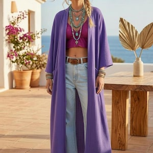 Könnte beinhalten: Ein langer, vorne offener, violetter Cardigan aus strukturiertem Stoff, getragen über einem magentafarbenen bauchfreien Oberteil und einer hellblauen Jeans mit braunem Ledergürtel. Das Outfit wird durch geschichtete türkisfarbene Ketten ergänzt.