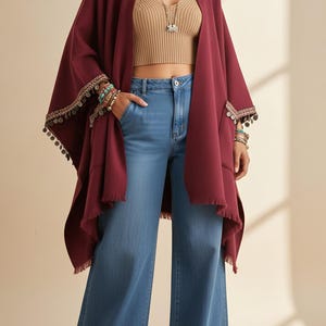 Può includere: Un poncho bordeaux con bordi sfrangiati e maniche decorate. Il poncho è abbinato a un top corto beige e a jeans blu chiaro a gamba larga. Le maniche sono decorate con piccole monete decorative.