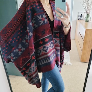 Könnte beinhalten: Ein ponchoartiger Wickel in Burgunderrot, Marineblau und Grau mit Muster. Der Wickel hat geometrische und Stammesmuster. Das Kleidungsstück ist vorne offen und hat weite Ärmel. Die Person trägt eine blaue Jeans und ein schwarzes Oberteil.
