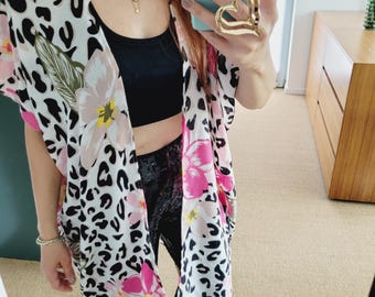 Pode incluir: Quimono branco com estampa de leopardo preto e detalhes florais rosa. O quimono é combinado com calças pretas e um anel dourado em forma de coração. Uma blusa preta e colares dourados também são visíveis.
