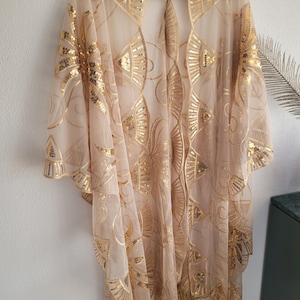 May include: A sheer beige kimono featuring an intricate gold sequin pattern of fan-like shapes and swirling lines. The garment has an open front, wide sleeves, and a hemline finished with long, thin gold fringe tassels hanging from the bottom edge.