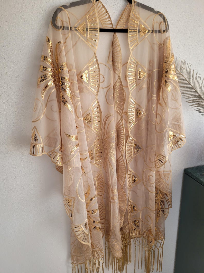 May include: A sheer beige kimono featuring an intricate gold sequin pattern of fan-like shapes and swirling lines. The garment has an open front, wide sleeves, and a hemline finished with long, thin gold fringe tassels hanging from the bottom edge.