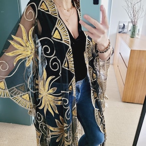 May include: A sheer black kimono featuring intricate gold sequin embroidery in floral and geometric patterns. The garment has wide, flowing sleeves, an open front design, and a hemline finished with long, thin black fringe detailing.