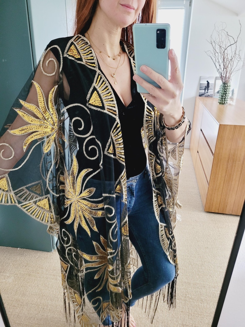 May include: A sheer black kimono featuring intricate gold sequin embroidery in floral and geometric patterns. The garment has wide, flowing sleeves, an open front design, and a hemline finished with long, thin black fringe detailing.