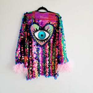 Könnte beinhalten: Eine sequinenbesetzte Jacke mit einem regenbogenfarbenen, schillernden Finish. Die Jacke hat lange Ärmel mit rosa Federbesatz an den Ärmelbündchen. Die Rückseite der Jacke zeigt ein großes pinkfarbenes Herz mit einem blauen Auge in der Mitte.