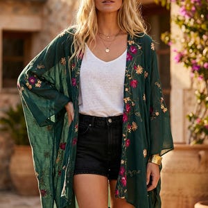 May include: A woman wearing a dark green kimono-style cover-up with floral embroidery in pink, yellow, and gold. She is also wearing a white top, black shorts, and gold jewelry. The background features a stone building and potted plants.