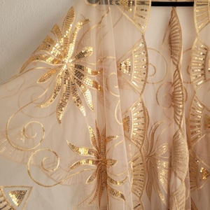 Könnte beinhalten: Ein durchsichtiger, beigefarbener Schal mit aufwendigen goldenen Pailletten- und Stickmustern mit Blumen- und geometrischen Motiven. Der Schal hängt an einem schwarzen Kleiderbügel und zeigt den zarten Stoff und die glitzernden Verzierungen. Das Design erinnert an den Art-Deco-Stil.