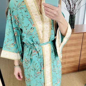Puede incluir: Un kimono azul turquesa con estampado floral y ribete dorado. El kimono tiene un cinturón de atar y mangas largas.