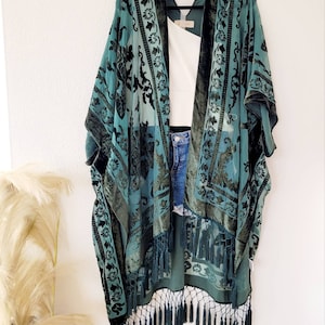 Puede incluir: Un kimono de terciopelo azul verdoso con un patrón floral negro y ribete de flecos. El kimono está colgado en una percha contra una pared blanca.