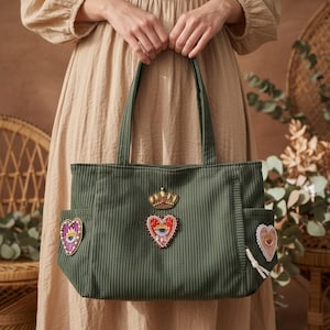 Può includere: Borsa tote in velluto a coste verde con toppe decorative. La borsa ha due tasche esterne e due manici. Le toppe includono una corona d'oro, un cuore con un occhio e un cuore con un motivo floreale.