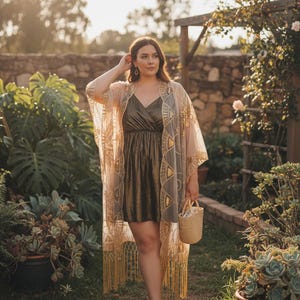 May include: A sheer, beige kimono featuring intricate gold embroidery and long fringe at the hem, worn over a metallic bronze, sleeveless mini dress with a V-neckline. A small, light-coloured woven straw handbag is held in one hand.