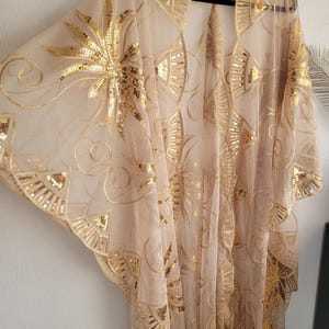 May include: A sheer beige kimono-style cover-up featuring intricate gold sequin embroidery in fan and floral patterns. The garment has wide, flowing sleeves, an open front design, and a decorative fringe trim along the bottom hemline.