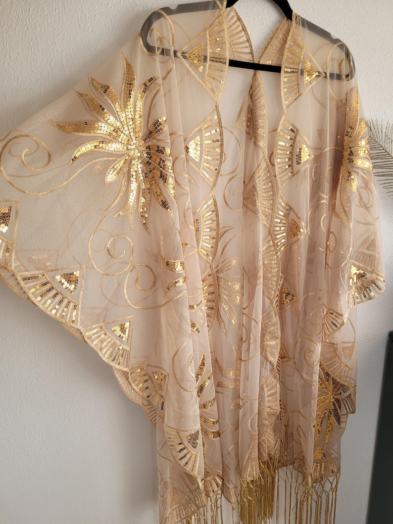May include: A sheer beige kimono-style cover-up featuring intricate gold sequin embroidery in fan and floral patterns. The garment has wide, flowing sleeves, an open front design, and a decorative fringe trim along the bottom hemline.