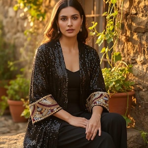 Könnte beinhalten: Eine Frau trägt eine schwarze Paillettenjacke mit aufwendig bestickten Manschetten, kombiniert mit einer schwarzen Hose. Die Jacke glitzert im Sonnenlicht. Der Hintergrund zeigt eine Steinmauer und Topfpflanzen, was einen raffinierten, eleganten Look erzeugt.