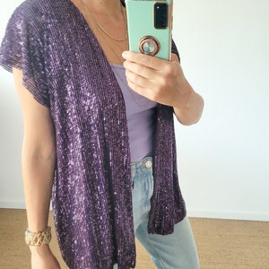 May include: A purple sequined cardigan with short sleeves. The cardigan is open in the front and is being worn over a purple tank top and blue jeans.