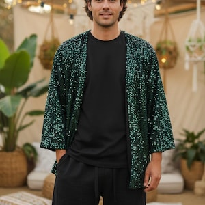 Könnte beinhalten: Eine Person trägt ein schwarzes T-Shirt und eine schwarze Hose mit einer grün glitzernden Jacke. Die Jacke hat eine lockere Passform und Dreiviertelärmel. Der Hintergrund umfasst Pflanzen und dekorative Lichter.