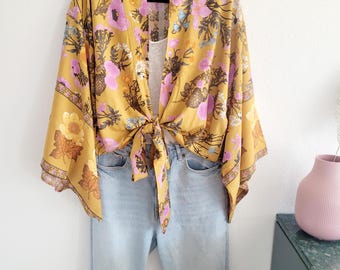 Pode incluir: Quimono floral amarelo mostarda com detalhes de flores rosa e roxas, amarrado na cintura. Jeans azul claro são exibidos abaixo do quimono. O quimono está pendurado em um cabide preto contra uma parede branca.
