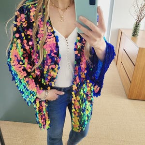 Puede incluir: Una mujer lleva una chaqueta larga de lentejuelas iridiscentes con ribete azul. La chaqueta tiene un efecto arcoíris con lentejuelas rosas, verdes y azules. Lleva pantalones vaqueros azules y está descalza.