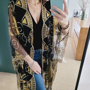 May include: A black sheer kimono featuring intricate gold sequin embroidery in geometric fan and swirl patterns. The garment has an open front, wide sleeves, and a fringe hemline. It is worn over a black top and blue denim jeans.