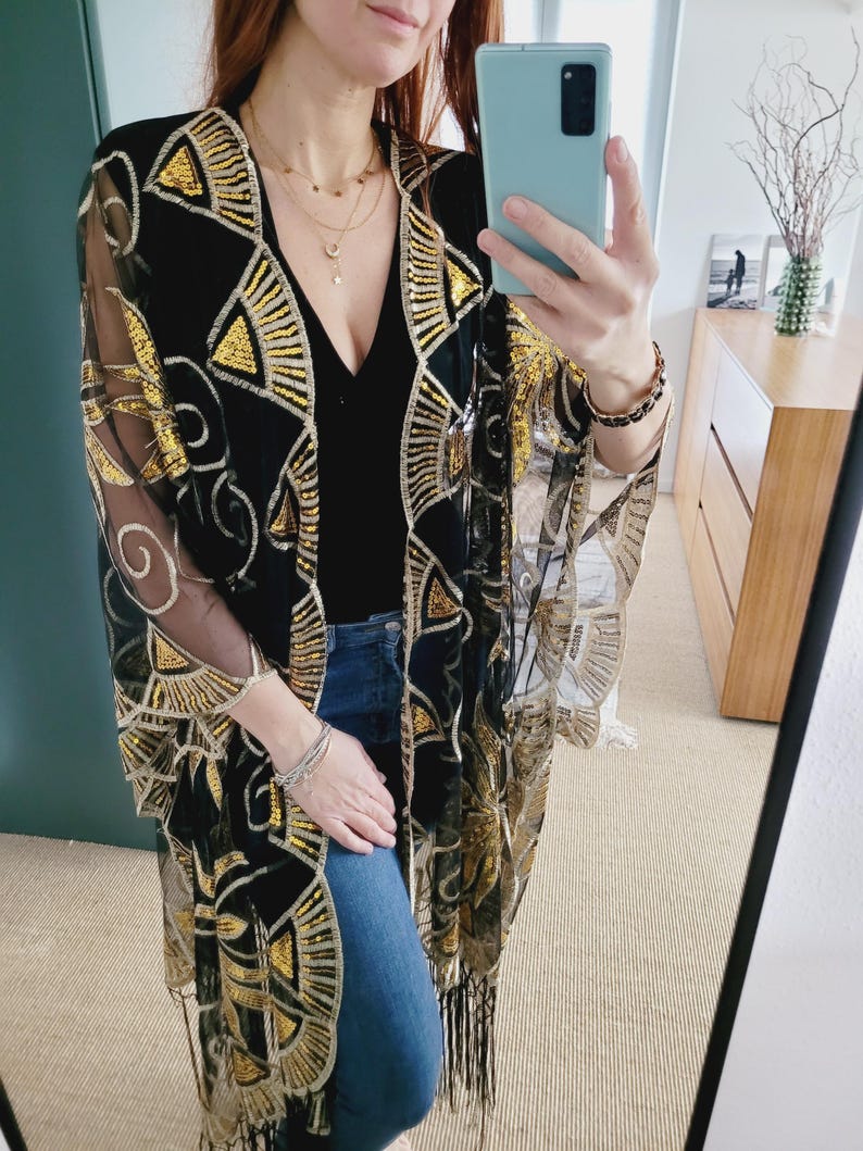 May include: A black sheer kimono featuring intricate gold sequin embroidery in geometric fan and swirl patterns. The garment has an open front, wide sleeves, and a fringe hemline. It is worn over a black top and blue denim jeans.