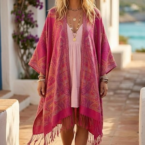 Peut inclure: Une femme porte un châle rose à motifs cachemire et franges, sur une robe rose clair. Elle porte plusieurs colliers et bracelets dorés. La scène se déroule sur un chemin pavé, avec une vue côtière en arrière-plan.