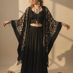 Peut inclure: Un ensemble noir composé d'une jupe longue et fluide et d'un haut court en dentelle, associé à un châle noir ouvert orné de broderies florales complexes dans des tons bleu, blanc et doré, terminé par des franges le long de l'ourlet.
