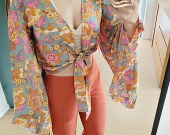 Pode incluir: Um top envolvente com estampa floral com mangas sino, amarrado na cintura, combinado com calças cor de ferrugem. A parte superior apresenta um fundo azul claro com estampas florais laranja, rosa e amarelo. Um anel dourado em forma de coração é visível.