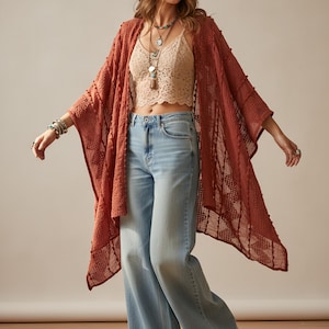 Puede incluir: Una mujer con un kimono abierto de color óxido con un patrón texturizado. También lleva un top corto de encaje beige, vaqueros de pernera ancha azul claro y collares superpuestos. El kimono tiene un diseño suelto y fluido.