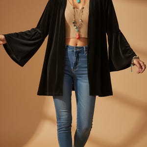 Op de afbeelding: Een zwarte fluwelen jas met open voorkant en wijde klokmouwen. De jas wordt gecombineerd met een geribbelde crop top en een blauwe jeans. Het model draagt gelaagde kettingen en armbanden. De achtergrond is effen beige.