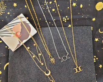 May include: Several gold and silver necklaces with celestial and symbolic pendants, including moons, stars, and an Ankh symbol. The necklaces are presented on a black surface with a box featuring an illustration. The background is a black fabric with gold star patterns.