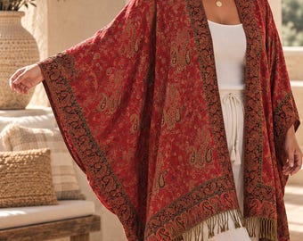 Pode incluir: Um quimono vermelho aberto na frente com um padrão paisley escuro, uma borda preta larga com detalhes intrincados em dourado e vermelho, e franjas longas verde-oliva na bainha inferior, usado sobre uma blusa e calças brancas.