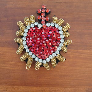 Könnte beinhalten: Ein herzförmiges Ornament mit einem roten Paillettenzentrum, umgeben von weißen und goldenen Perlen. Oben befindet sich ein kleines Kreuz. Das Design befindet sich auf einer schwarzen Basis.