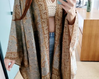 May include: A gray and brown paisley patterned kimono-style wrap with fringe. The wrap features a detailed design with a mix of gray and brown tones. The garment is worn over a white crochet top and blue jeans.
