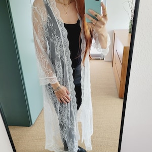 May include: A white lace long-sleeve cardigan with a delicate floral pattern. The cardigan is sheer and has a relaxed fit.