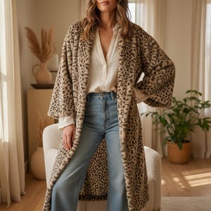 Könnte beinhalten: Ein langer, offener Mantel mit Leopardenmuster in Beige- und Brauntönen. Der Mantel hat weite Ärmel und eine weiche Kunstfellstruktur. Das Model trägt eine weiße Bluse und blaue Jeans.