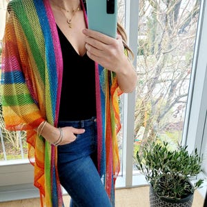Könnte beinhalten: Ein regenbogenfarbener gestrickter Kimono mit Fransen. Der Kimono wird über einem schwarzen Tanktop und einer blauen Jeans getragen.