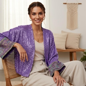 Könnte beinhalten: Eine Frau trägt einen lila Pailletten-Kimono mit bestickten Manschetten. Die Jacke ist offen und zeigt ein cremefarbenes Oberteil und helle Hosen. Die Frau sitzt auf einem Korbstuhl und lächelt in die Kamera.