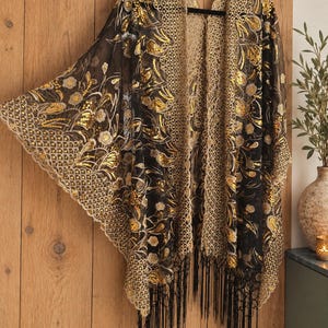 May include: A black sheer kimono featuring gold sequin floral embroidery, a wide scalloped lace border along the front opening and hem, and long black fringe trim at the bottom. The garment is displayed on a black hanger against a wooden wall.
