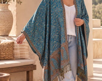 May include: A teal blue open-front kimono wrap featuring an intricate floral and paisley pattern in shades of brown and gold. The garment has a wide border along the front edges and hem, finished with long, dark olive green fringe along the bottom.