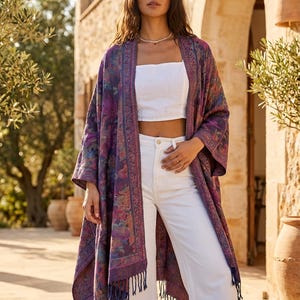 Può includere: Un lungo kimono aperto sul davanti, nei toni del viola, blu e rosa, con un motivo floreale e finiture a frange. Il kimono è indossato sopra un top corto bianco e pantaloni bianchi. Lo sfondo comprende un edificio in pietra e ulivi.
