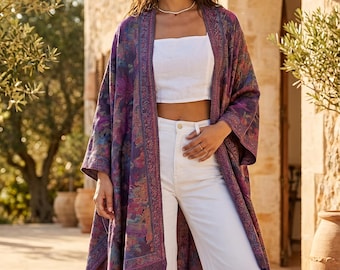 May include: A long, open-front kimono in shades of purple, blue, and pink, with a floral pattern and fringe trim. The kimono is worn over a white crop top and white trousers. The background includes a stone building and olive trees.