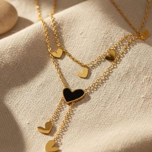 Könnte beinhalten: Eine goldfarbene Halskette mit einem mehrschichtigen Design mit herzförmigen Anhängern. Die Halskette hat einen zentralen schwarzen Herzanhänger und kleinere goldene Herzen. Der Schmuck wird auf einem beigefarbenen Stoff präsentiert.