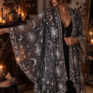 Puede incluir: Un kimono negro transparente con intrincados bordados de lentejuelas plateadas en patrones celestiales, incluyendo rayos de sol, lunas crecientes y estrellas. La prenda tiene mangas anchas y un dobladillo festoneado con un borde plateado.